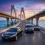 A high-resolution, square photograph capturing a powerful fleet of three glistening black ghtrips.com chauffeur SUVs, including a Cadillac Escalade and GMC Yukon Denali, moving in loose formation across the intricate modern bridge structure of the King Fahd Causeway towards Bahrain, under a dramatic golden-hour sky - ghtrips.com